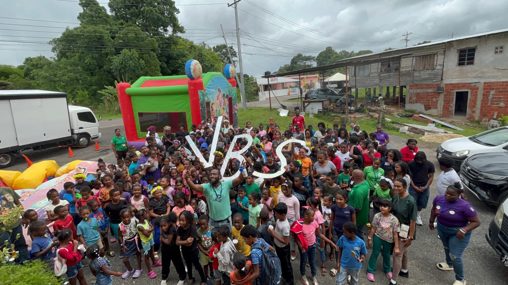 Large group photo of VBS 2025 attendees outdoors with bouncy house and VBS sign